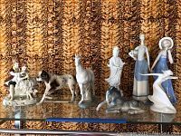 Ceramic figurines including two ladies in blue dresses with hats, Asian-themed figure, a white horse, spotted horse, reclining horse, spaniel dog, and a swan figurine, arranged on glass shelf.