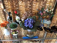 Full lot view of faux flowers, jeweled bonsai trees, glass squirrel figurine, and trinket bowls on glass shelf.