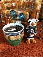 Planter with soil inside, two glass water orbs inserted, standing next to a 16-inch winter-themed penguin figurine, all placed on orange carpet with plaid patterned chair in background.