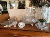 Wide shot of multiple crystal items grouped including shoe, clock, pitcher, vase, bowl, and trays on wooden surface