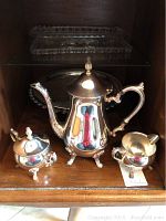 Silver plated tea pot, creamer, sugar bowl arranged on wooden shelf with a large round serving platter and cut glass platter behind