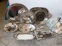 Full view of all silverplate serving items grouped on floor including platters, bowls, bread basket, and utensils with dust bags netted behind