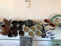 Multiple ceramic oriental tea sets including teapots, cups, fish chopstick rests, bird plate, and mugs arranged on glass shelf.