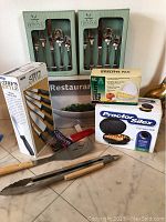 Photo shows boxed cutlery sets, Chef's Style knife set, compact grill, omelet pan, grill accessories, and serving dish box.