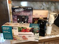 Three kitchen appliances boxed and unboxed on marble countertop: DeLonghi Caffé Setta espresso and cappuccino maker box with frother, Freshlock vacuum sealer box, and Kitchen Magician appliance.