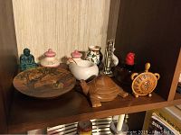 Shelf with various international figurines including a green ceramic Buddha, wooden turtle, ceramic jars with lids, brass candlestick, and ship's wheel ornament.