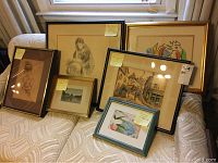 Seven framed artworks displayed on a couch including sketches, watercolor, print, photograph, and textile image