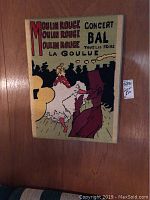 Needlepoint reproduction of the Moulin Rouge poster featuring a dancing figure in yellow, red, and black tones on beige background hung on wooden wall.
