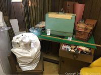 Overview of green table with vintage Royal sewing machine accessories including a green hard case, sewing supplies in drawer, fabric pieces, and a large white bag beside the table.