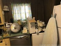 Photo showing Waring stand mixer with yellow base and glass bowl, chrome electric fan, blender, toaster, iron, and espresso maker on kitchen counters against wood paneled wall and curtained window.