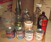 Photo showing a collection of six advertising tins, glass bottles of various sizes, and a metal trash can with visible rust.