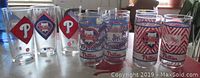 Front view of eleven Philadelphia Phillies promotional glasses with various logos and decals, displayed on table with light background.
