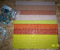 Eight sheets of glass tiles arranged by color on wooden surface, with two bags of loose mosaic pieces and black and white ceramic tiles scattered nearby.