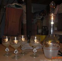 Photo shows the Bohemian glass decanter alongside five stemmed glasses on a table in a storage space, highlighting the gold leaf and frosted decoration.