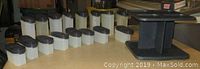Photo showing all 16 white rectangular Tupperware spice containers with black lids lined up on a table along with the black rotating rack in the background.