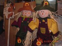 Photo of two scarecrows standing side by side wearing colorful autumn clothes with straw hair and hats with sunflowers, cardboard background with Halloween signs.