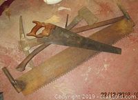 Photo showing four vintage hand tools: 2-man hand saw, hand saw, pick, and axe laid out on a surface, noting wooden handles and rust on metal parts