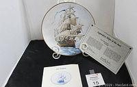 Plate displayed upright on a stand alongside Certificate of Authenticity and informational booklet against a black background.