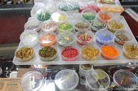 Plastic display case holding 18 clear plastic jars filled with glass beads and bugles in various colors including gold, green, orange, red, blue.