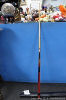 Full length view of the pool stick with natural top and black lower section featuring painted roses, leaning against a blue backdrop with toys on top of the background.