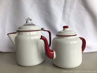 Front view of both vintage enamelware pieces showing white bodies with red handles and lids, one taller than the other