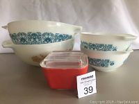Overall lot showing 2 large white glass mixing bowls with different vintage floral patterns stacked, 2 small bowls with blue pattern stacked, and 1 small red covered dish in front.
