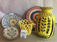 Front view of five Italian ceramics on white surface showing a tall yellow vase with black floral designs, three hand-painted floral plates, and a yellow lattice-style basket.