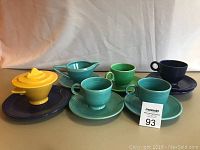 Photo showing 4 cups in turquoise, green, navy blue colors, 5 matching saucers, yellow covered sugar bowl on navy blue plate, small pitcher, and one additional plate.