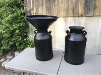 Photo showing both antique black milk cans side by side on a table outdoors, one with a wide removable sifter funnel top, the other without a top and with one loose handle end.