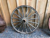 Full view of the antique wooden wagon wheel leaning against a wooden fence on gravel ground, showing the entire wheel structure and original black paint on the spokes.