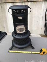 Front view of the black metal antique oil heater showing the full height and base with perforated design. Measuring tape shows height about 22 inches.