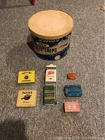 Photo of 6 small vintage advertising tins arranged in two rows on carpeted floor with a large Old Swiss Potato Chips cardboard container behind them. A tape measure on the floor indicates scale.