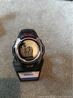 Close-up image of the Casio G-Shock digital watch displaying date and time, mounted on a branded stand.
