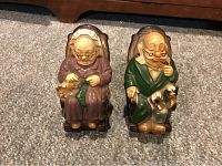 Pair of vintage ceramic grandparents retirement fund piggy banks shown seated, front view on carpeted floor.