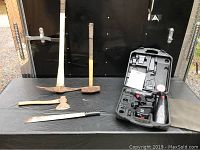 Full view of the five tools arranged on table against black background: pickaxe, sledgehammer, machete, small axe, and open case with battery drill and accessories.