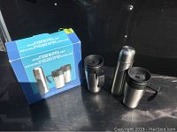 Photo of the stainless steel thermos and two coffee mugs with black lids and handles, with their original box in background.