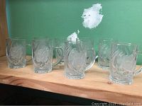 Photo showing 8 clear glass mugs with frosted flower designs arranged on a wooden surface against a green wall.