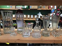Front view showing assortment of clear glass vases, decanter, drinking glasses, and candle stick holder arranged on a wooden shelf.