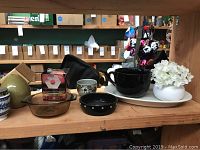 Photo showing various ceramics including a large black ceramic bowl, black ashtray, tan decanter, hexagon game box, white ceramic vase with flowers, and large white oval platter on wooden shelf.