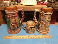 Three ceramic steins displayed on a blue surface with a ruler for size reference, showing California, country scene, and Louisiana Bourbon Street designs.