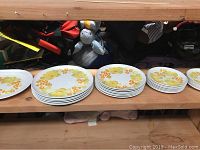 Photo showing stacks of plates lined up on a shelf, organized by type, with visible yellow and orange floral decoration on white background.