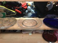 Three glass cake plates: one large clear plate, one clear plate with beaded edge, and one blue glass pedestal plate displayed on a wooden surface.