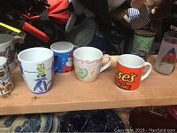 Nine assorted mugs and glasses on wooden surface, various themed prints visible