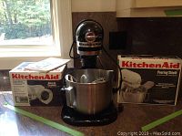 Black KitchenAid heavy duty stand mixer with stainless steel mixing bowl on countertop with boxed slicer & shredder attachment and boxed pouring shield
