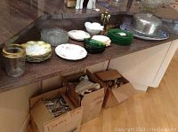 Kitchen countertop with stacked vintage glass plates and dishes, brass candleholder, aluminum cake cover, and cardboard boxes below