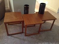Three wooden end tables arranged side by side fitting together like puzzle pieces, medium brown wood finish, tables approximately 18x16x16.5 inches each. Two metallic finish floor vases positioned behind the tables.