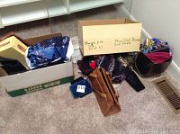 Two cardboard boxes on floor containing assorted scarves, clutches, and fabric items, along with folded nylon cart and plastic poncho partly visible.