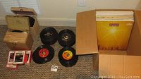 Overview photo showing vinyl 45 RPM records lying on floor near boxes containing eight-track tapes and LP albums.