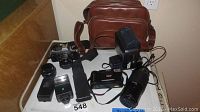 Overview photo of all the lot items displayed on a table including cameras, lenses, flashes, viewcam recorder, and leather bag.