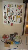 Photo of pegboard with hanging tools, hardware packages, and cords; hand axe, hammer, pliers, tape measures, and fasteners visible. Shop vac and drill on floor below.